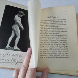 May include: Open book with a black and white image of a nude male figure. The page on the left has handwritten text and a signature. The right page has printed text titled "PREFACE."