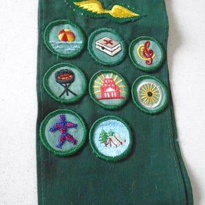 May include: A dark green sash with embroidered patches. Patches include a beach scene, first aid kit, musical note, barbecue grill, lighthouse, wagon wheel, abstract figure, and a campsite. A yellow and green wing emblem is at the top.