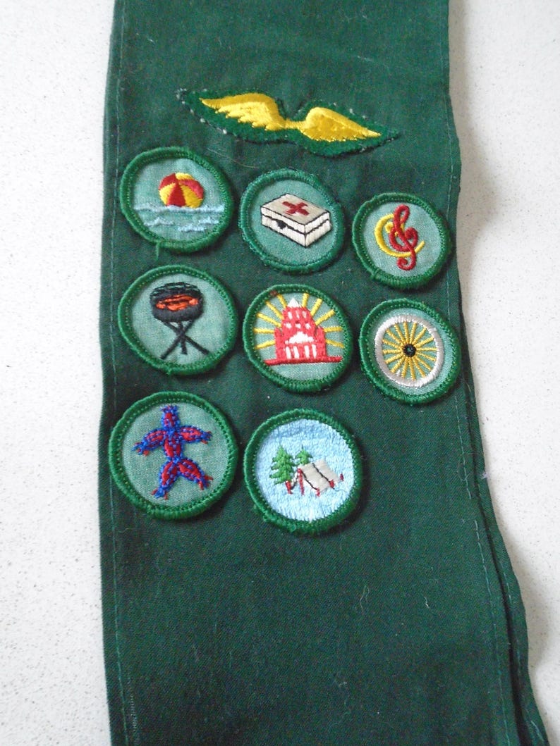 May include: A dark green sash with embroidered patches. Patches include a beach scene, first aid kit, musical note, barbecue grill, lighthouse, wagon wheel, abstract figure, and a campsite. A yellow and green wing emblem is at the top.