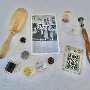 May include: A collection of vintage sewing supplies including a wooden mallet, a spool of black thread, a card of pearl buttons, a thimble, a vintage photo, and a small wooden tool with a wheel.