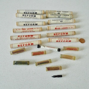 May include: An assortment of vintage pencil lead refills and accessories. The image features several cylindrical paper tubes labeled "REFORM" and "EBERHARD FABER", along with small plastic containers of colored leads and a black pen nib.
