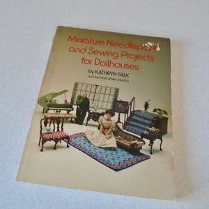 May include: A vintage book titled "Miniature Needlepoint and Sewing Projects for Dollhouses" by Kathryn Falk. The cover features a colorful illustration of miniature dollhouse furniture and figures, including a piano, chairs, and a sofa.
