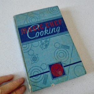 May include: Vintage "Magic Chef Cooking" cookbook. The cover is light blue with a dark blue banner containing the title in red and white. Decorative circles and lines adorn the cover. A red and blue emblem is in the lower right corner.