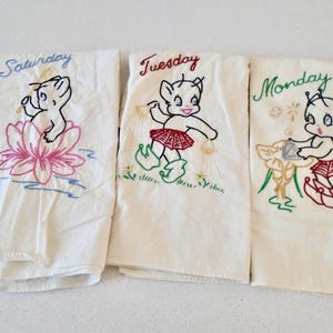 May include: Three white tea towels with embroidered images of cartoon characters and the words "Saturday", "Tuesday", and "Monday".