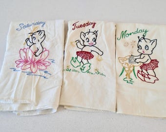 May include: Three white tea towels with embroidered images of cartoon characters and the words "Saturday", "Tuesday", and "Monday".