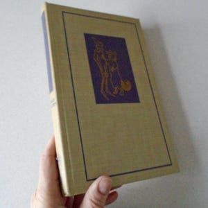 May include: A vintage hardcover book with a tan cover and a purple illustration of two figures on the front. The illustration is framed with a black border.
