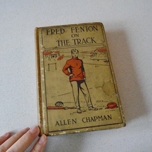 May include: Vintage book titled "Fred Fenton on the Track" by Allen Chapman. The cover features an illustration of a young man in an orange shirt and gray pants. The book has a worn, aged appearance, with a tan cover and visible wear.