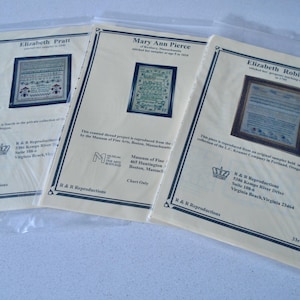 May include: Three packaged cross-stitch sampler charts. Each chart features a framed sampler design with text. The charts are labeled "Elizabeth Pratt", "Mary Ann Pierce", and "Elizabeth Robinson". The charts are in plastic sleeves.