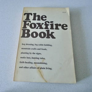 May include: A softcover book titled The Foxfire Book with a cream-colored cover and dark brown text. The cover lists topics including hog dressing, log cabin building, mountain crafts, foods, planting by the signs, snake lore, hunting tales, faith healing, and moonshining.