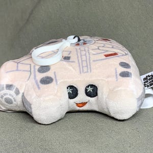 Puede incluir: Juguete de peluche blanco con forma de Halcón Milenario de Star Wars. El juguete tiene un lazo blanco para colgar y una etiqueta con el logotipo de Star Wars.