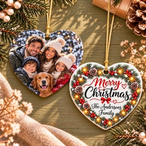 Puede incluir: Dos adornos navideños de cerámica en forma de corazón con cordones dorados. Uno muestra una foto familiar con un perro, el otro una corona de ramas de pino, bayas rojas y luces con el texto Merry Christmas The Andersons Family.