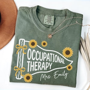 Puede incluir: Una camiseta verde oliva con un gráfico de banderín blanco con el texto "OCCUPATIONAL THERAPY" y "Mrs. Emily" en letra blanca. El diseño incluye girasoles amarillos y dos lazos amarillos en el lado izquierdo del banderín.