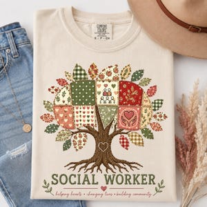 Puede incluir: Una camiseta de color crema con el gráfico de un árbol con una copa de patchwork hecha de varios patrones que incluyen corazones, flores y un icono comunitario. El texto SOCIAL WORKER está impreso debajo del árbol con la frase helping hearts, changing lives, building community.