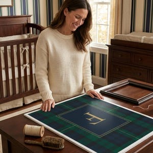 May include: A framed plaid print with a gold letter "F" on a navy blue background. The print is surrounded by a white border. A spool of thread and a brush are on the table. A woman is looking at the print.