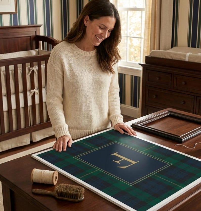 May include: A framed plaid print with a gold letter "F" on a navy blue background. The print is surrounded by a white border. A spool of thread and a brush are on the table. A woman is looking at the print.