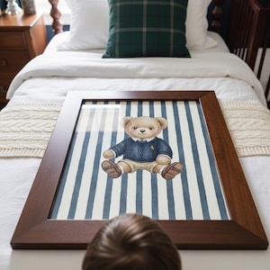 May include: A framed print featuring a watercolor illustration of a teddy bear wearing a blue sweater and khaki shorts, set against a backdrop of blue and white vertical stripes. The frame is a dark brown wood.