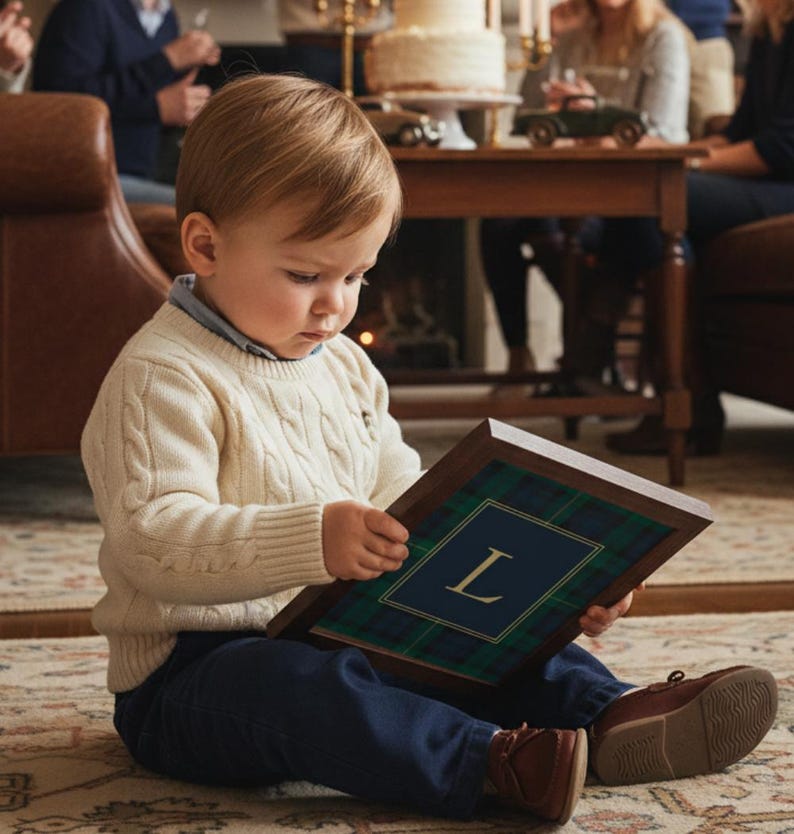 May include: A young child wearing a cream-colored cable knit sweater and navy blue pants sits on a patterned rug, holding a framed print. The print features a blue background with the letter "L" and a green and blue plaid border.