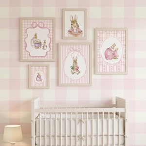 May include: A set of framed watercolour prints depicting rabbits in charming scenes. The artwork is displayed on a pink and white checked wall, with a white cot and a lamp in the foreground. The frames are a neutral colour.