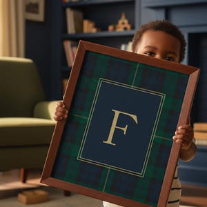 May include: A framed print featuring a navy blue and gold letter "F" on a navy blue background, surrounded by a green and navy plaid pattern. The frame is a dark brown color. The print is held by a person.