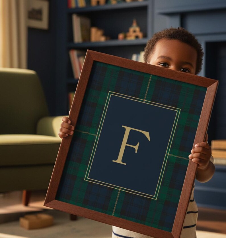 May include: A framed print featuring a navy blue and gold letter "F" on a navy blue background, surrounded by a green and navy plaid pattern. The frame is a dark brown color. The print is held by a person.