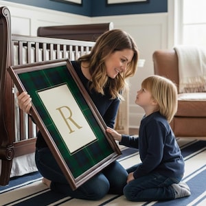 May include: A framed artwork featuring a green and navy plaid pattern with a white center and a gold letter "R". The artwork is held by a person kneeling on a striped rug, looking at a child. A dark wood crib is in the background.
