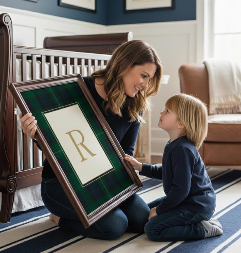 May include: A framed artwork featuring a green and navy plaid pattern with a white center and a gold letter "R". The artwork is held by a person kneeling on a striped rug, looking at a child. A dark wood crib is in the background.