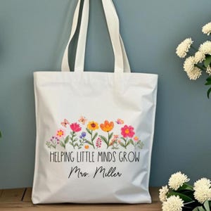 Op de afbeelding: Witte draagtas met een bloemenontwerp en de tekst "Helping Little Minds Grow Mrs. Miller." Het ontwerp bevat kleurrijke bloemen en vlinders. De tas heeft twee witte handvatten.