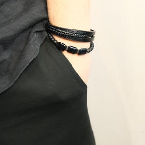 May include: Two black bracelets are worn on the wrist. One is a braided leather band, and the other is a beaded bracelet with rectangular black beads. The bracelets are displayed against a black shirt and a light background.