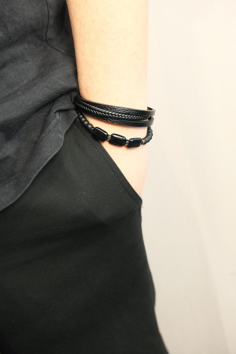 May include: Two black bracelets are worn on the wrist. One is a braided leather band, and the other is a beaded bracelet with rectangular black beads. The bracelets are displayed against a black shirt and a light background.