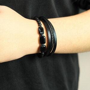 May include: Black beaded bracelet with three rectangular black stones, paired with a braided leather bracelet and a simple leather band. The bracelets are worn on a person's wrist, against a black shirt.