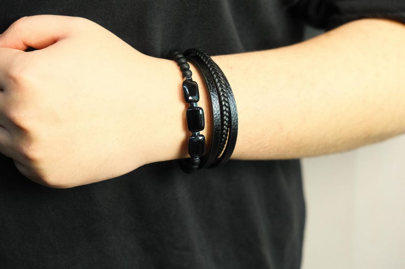 May include: Black beaded bracelet with three rectangular black stones, paired with a braided leather bracelet and a simple leather band. The bracelets are worn on a person's wrist, against a black shirt.