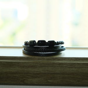May include: A stack of black bracelets with various designs. The bracelets include beaded, braided, and leather styles. The bracelets are displayed on a wooden surface with a blurred background.