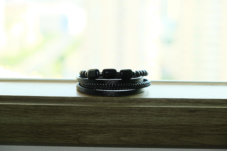 May include: A stack of black bracelets with various designs. The bracelets include beaded, braided, and leather styles. The bracelets are displayed on a wooden surface with a blurred background.