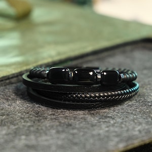 May include: A black bracelet with multiple strands. The top strand has black beads and three rectangular black stones. Below are leather strands, including a braided one. The bracelet is set against a grey surface.