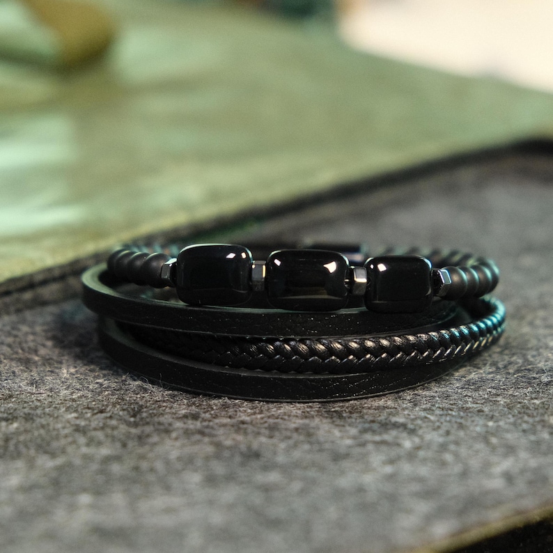 May include: A black bracelet with multiple strands. The top strand features black beads and three rectangular black stones. Below are leather strands, including a braided one. The bracelet is set against a gray surface.