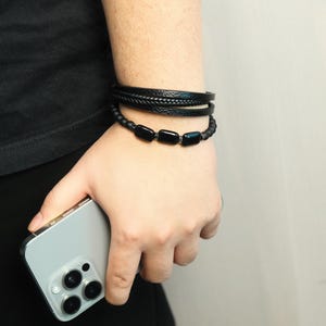 May include: A person's wrist is adorned with three black bracelets. The top two bracelets are leather, one braided and the other smooth. The bottom bracelet features black rectangular beads. A silver smartphone is held in the hand.