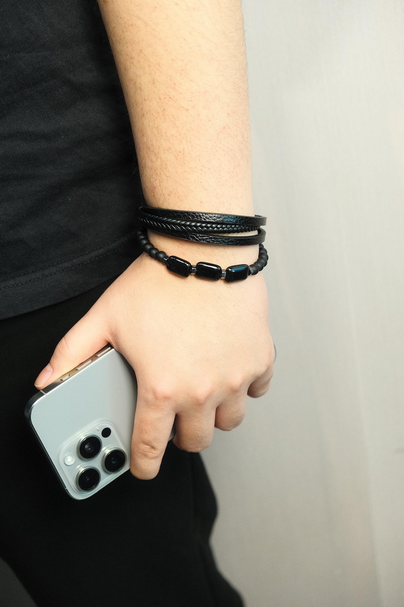 May include: A person's wrist is adorned with three black bracelets. The top two bracelets are leather, one braided and the other smooth. The bottom bracelet features black rectangular beads. A silver smartphone is held in the hand.