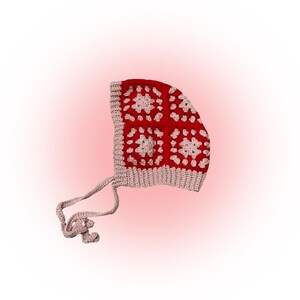 May include: Red and white crocheted baby bonnet with a white tie. The bonnet is made with a granny square pattern.