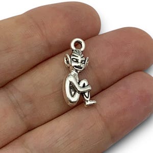 May include: A silver-coloured charm featuring a seated goblin or elf with pointed ears and a grin. The charm has a loop at the top for attaching to a necklace or bracelet. The goblin is sitting with its knees up, and its arms are wrapped around its legs.