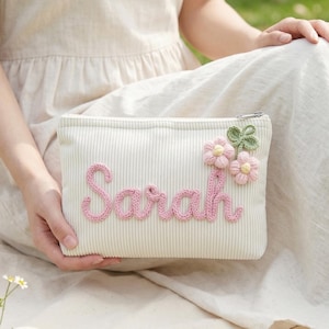 Può includere: Un astuccio in velluto a coste color crema con il nome "Sarah" ricamato in rosa. È decorato con fiori rosa e verdi. Lo sfondo include un cestino da picnic, un libro e dei fiori.