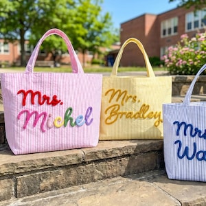 May include: Three seersucker tote bags in pink, yellow, and blue stripes. The pink bag features the text Mrs. Michel in multi-colored yarn. The yellow bag displays the text Mrs. Bradley in gold yarn, and the blue bag shows Mrs. Walker in blue.