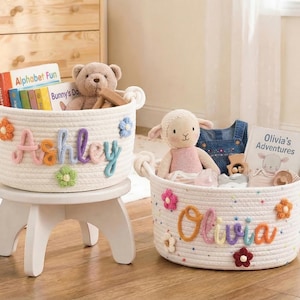 Puede incluir: Dos cestas de cuerda blanca con asas, decoradas con flores coloridas y nombres. Una cesta dice "Ashley" y está sobre un taburete blanco, llena de libros y un osito de peluche. La otra dice "Olivia" y contiene un peluche de cordero y un libro.