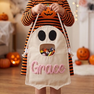May include: A cream-colored canvas trick-or-treat bag shaped like a ghost with two black felt eyes and a large open mouth filled with candy. The name Grace is embroidered in pink yarn across the bottom. The bag features two white rope handles.