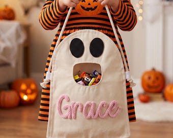Op de afbeelding: Een crèmekleurige canvas tas voor snoep in de vorm van een spook met twee zwarte vilten ogen en een grote open mond gevuld met snoepjes. De naam Grace is onderaan geborduurd in roze garen. De tas heeft twee witte touw-handvatten.