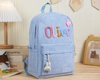 May include: Pale blue corduroy backpack with the name "Olive" in colourful yarn letters. Features a small pink flower and a white bunny charm. Includes a front pocket and side pocket.