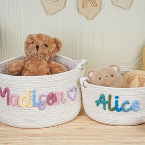 May include: Two white rope baskets with handles, one larger and one smaller. The larger basket has the name "Madison" in colorful letters, and a teddy bear inside. The smaller basket has the name "Alice" and a small teddy bear.