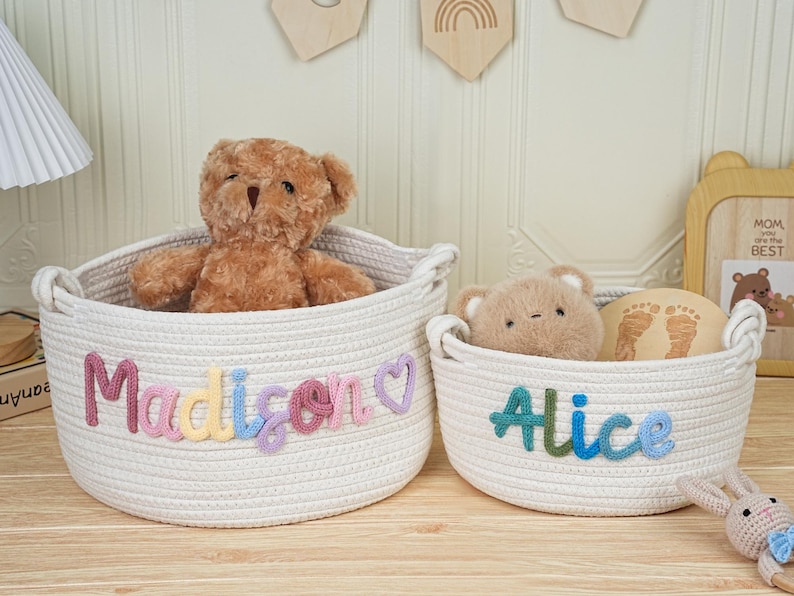 May include: Two white rope baskets with handles, one larger and one smaller. The larger basket has the name "Madison" in colorful letters, and a teddy bear inside. The smaller basket has the name "Alice" and a small teddy bear.