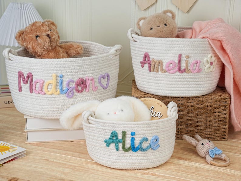 May include: Three white rope baskets with embroidered names: "Madison", "Amelia", and "Alice". Each basket contains a stuffed animal. The "Madison" basket has a teddy bear, the "Amelia" basket has a teddy bear and a pink blanket, and the "Alice" basket has a stuffed bunny.