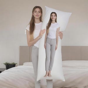 May include: A long, white body pillow held by a person. The pillow is rectangular and appears to be made of a soft material. The person is standing in front of a bed with a white duvet and a neutral-coloured headboard.