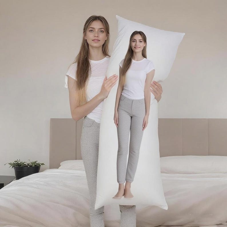 May include: A long, white body pillow held by a person. The pillow is rectangular and appears to be made of a soft material. The person is standing in front of a bed with a white duvet and a neutral-coloured headboard.